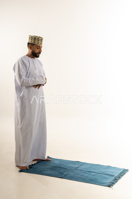 التوجه للقبلة وأداء فريضة الصلاة، التقرب إلى الله بالعبادة والطاعة، بورتريه لرجل عربي خليجي عماني يرتدي الدشداشة والكمه يقف على السجادة ويصلي، الحرص على عدم تفويت وقت الصلاة، الجسم بالطول الكامل، خلفية بيضاء