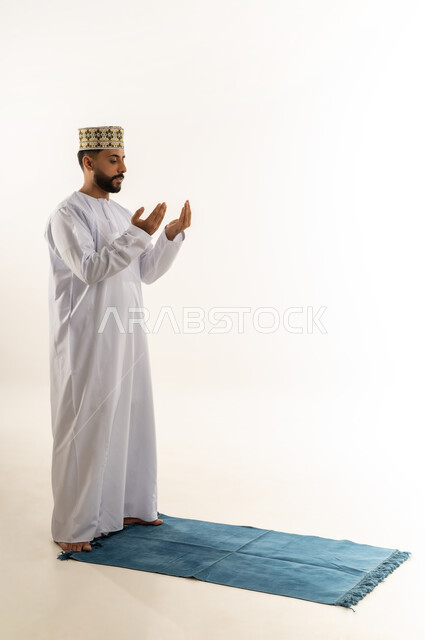 الخشوع والتضرع في الدعاء، التقرب إلى الله بالعبادة والطاعة، طلب المغفرة والعفو من الله، بورتريه لرجل عربي خليجي عماني يرتدي الدشداشة والكمه يقف على سجادة الصلاة يرفع كفيه المفتوحتين وينظر إلى الأعلى ويدعو بتذلل، الجسم بالطول الكامل، خلفية بيضاء