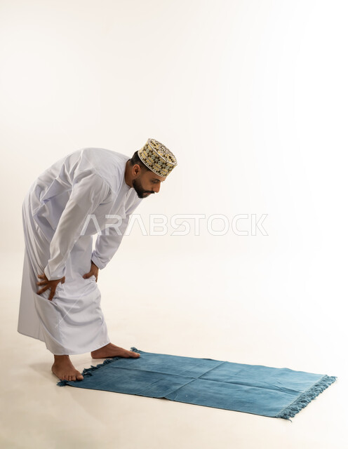 Kneeling and performing the obligatory prayer, being careful not to miss the prayer time, getting closer to God through worship and obedience, portrait of an Arab Gulf Omani man wearing a dishdasha and a cloak standing on the carpet and kneeling, white background