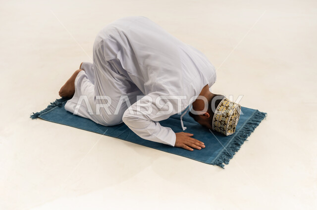 Prostrating on the carpet and performing the prayer on time, humility and supplication to God, portrait of an Arab Gulf Omani man wearing a dishdasha and a cloak performing the obligatory prayer, drawing closer to God through worship and obedience, white background