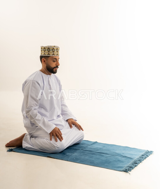 أداء الصلاة في وقتها، الجلوس على السجادة قبل التسليم، الخشوع والتضرع إلى الله، بورتريه لرجل عربي خليجي عماني يرتدي الدشداشة والكمه يؤدي فريضة الصلاة، التقرب إلى الله بالعبادة والطاعة، خلفية بيضاء