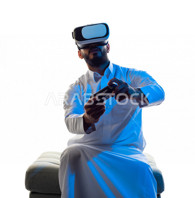 Portrait of a Saudi Arabian Gulf man wearing 3D virtual reality glasses, holding a joystick in his hand and playing video games with gestures that indicate focus, fun and entertainment in virtual world games, PlayStation entertainment games, techniques an