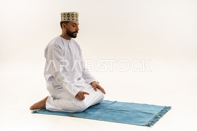 الجلوس للتشهد على السجادة، الخشوع والتضرع إلى الله، بورتريه لرجل عربي خليجي عماني يرتدي الدشداشة والكمه يؤدي فريضة الصلاة، أداء الصلاة في وقتها، التقرب إلى الله بالعبادة والطاعة، خلفية بيضاء