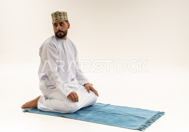 الجلوس على السجادة والتسليم، أداء الصلاة في وقتها، الخشوع والتضرع إلى الله، بورتريه لرجل عربي خليجي عماني يرتدي الدشداشة والكمه يؤدي فريضة الصلاة، التقرب إلى الله بالعبادة والطاعة، خلفية بيضاء