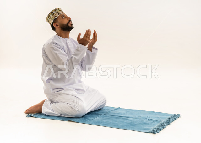 طلب المغفرة والعفو من الله، الدعاء بخشوع وتضرع، بورتريه لرجل عربي خليجي عماني يرتدي الدشداشة والكمه يجلس على سجادة الصلاة يرفع كفيه المفتوحتين للأعلى ويدعو بتذلل، التقرب إلى الله بالعبادة والطاعة، خلفية بيضاء