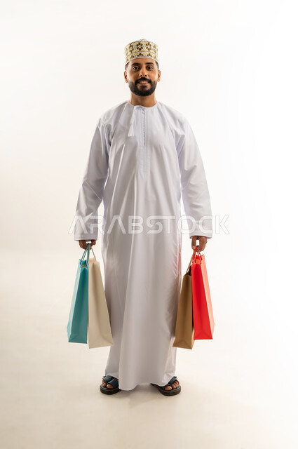اقتناء الهدايا والحاجات الضرورية، الاستفادة من العرض والخصم في المول، قضاء وقت ممتع في شراء المستلزمات، بورتريه لشاب عربي خليجي عماني مبتسم يرتدي الدشداشة والكمه يحمل أكياس التسوق الملونة، الأناقة والاهتمام بالمظهر الخارجي، الجسم بالطول الكامل، خلفية بيضاء