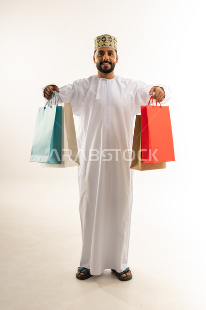 قضاء وقت ممتع في شراء المستلزمات، الاستفادة من العرض والخصم في المول، اقتناء الهدايا والحاجات الضرورية، بورتريه لشاب عربي خليجي عماني مبتسم يرتدي الدشداشة والكمه يحمل أكياس التسوق الملونة، الأناقة والاهتمام بالمظهر الخارجي، الجسم بالطول الكامل، خلفية بيضاء