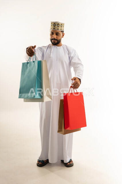 الاستفادة من العرض والخصم في المول، قضاء وقت ممتع في شراء المستلزمات، اقتناء الهدايا والحاجات الضرورية، بورتريه لشاب عربي خليجي عماني مبتسم يرتدي الدشداشة والكمه يحمل أكياس التسوق الملونة، الأناقة والاهتمام بالمظهر الخارجي، الجسم بالطول الكامل، خلفية بيضاء