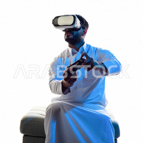 Portrait of a Saudi Arabian Gulf man wearing 3D virtual reality glasses, holding a joystick in his hand and playing video games with gestures that indicate focus, fun and entertainment in virtual world games, PlayStation entertainment games, techniques an
