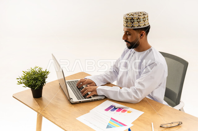 Using modern technologies in the office and administrative sector, the concept of work supervision, managing workflow via laptop, close-up portrait from the side of an Arab Gulf Omani man wearing a dishdasha and a cloak sitting behind a desk with a group of papers in front of him and a green plant next to him, white background