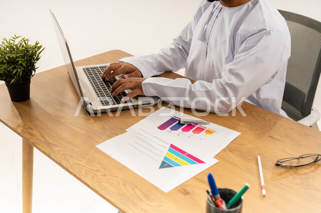 الكتابة على لوحة مفاتيح الحاسوب المحمول، الإشراف على العمل، استخدام التقنيات الحديثة في القطاع المكتبي والإداري، بورتريه مقرب من الجانب لرجل عربي خليجي عماني يرتدي الدشداشه يجلس خلف طاولة المكتب أمامه مجموعة مخططات واوراق وبجانبه نبتة خضراء، خلفية بيضاء