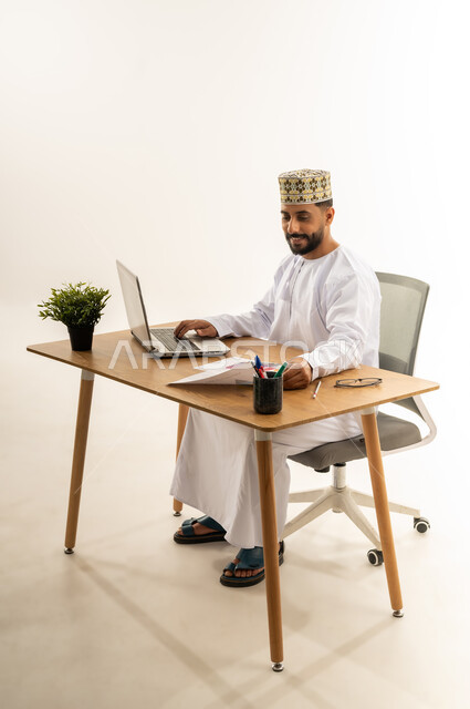 الاستفادة من التقنيات الحديثة في القطاع المكتبي والإداري، مفهوم الإشراف على العمل، إدارة سير الأعمال عبر الحاسوب المحمول، بورتريه من الجانب لرجل عربي خليجي عماني يرتدي الدشداشه والكمه يجلس خلف طاولة المكتب أمامه مجموعة أوراق وبجانبه نبتة خضراء، خلفية بيضاء