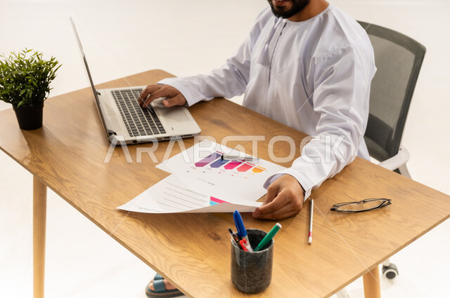 الإشراف على العمل، استخدام التقنيات الحديثة في القطاع المكتبي والإداري، إدارة سير الأعمال عبر الحاسوب المحمول، بورتريه مقرب من الجانب لرجل عربي خليجي عماني يرتدي الدشداشه يجلس خلف طاولة المكتب أمامه مجموعة مخططات واوراق وبجانبه نبتة خضراء، خلفية بيضاء