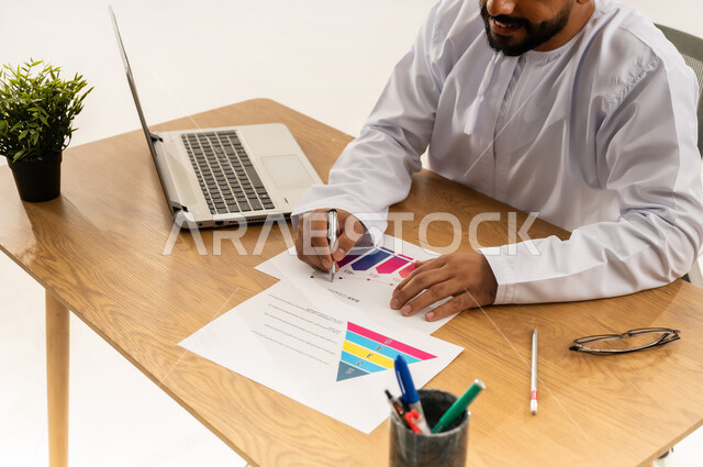 Reviewing and modifying the work plan and data, using modern technologies in the office and administrative sector, managing business via laptop, close-up portrait from the side of an Arab Gulf Omani man wearing a dishdasha sitting behind an office table with a group of papers in front of him and a green plant next to him, white background