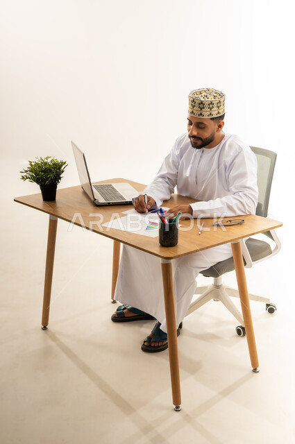 إدارة سير الأعمال عبر الحاسوب المحمول، مفهوم الإشراف على العمل، الاستفادة من التقنيات الحديثة في القطاع المكتبي والإداري، بورتريه من الجانب لرجل عربي خليجي عماني يرتدي الدشداشه والكمه يجلس خلف طاولة المكتب أمامه مجموعة أوراق وبجانبه نبتة خضراء، خلفية بيضاء