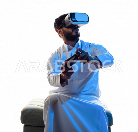 Portrait of a Saudi Arabian Gulf man wearing 3D virtual reality glasses, holding a joystick in his hand and playing video games with gestures that indicate focus, fun and entertainment in virtual world games, PlayStation entertainment games, techniques an