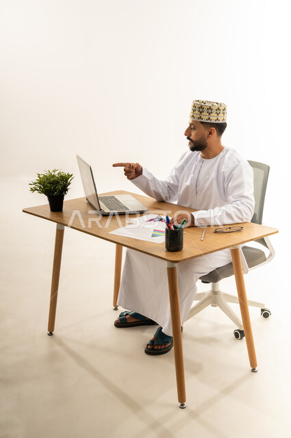 Pointing index finger at laptop screen, supervising work, taking advantage of modern technologies in the office and administrative sector, side portrait of an Arab Gulf Omani man wearing a dishdasha and a sleeve sitting behind an office table with a group of papers in front of him and a green plant next to him, white background