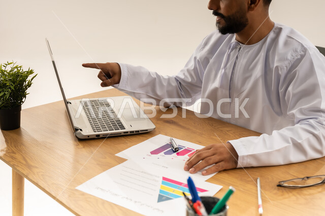 دمج التكنولوجيا والتقنيات الحديثة في القطاع المكتبي والإداري، إدارة الأعمال عبر الحاسوب المحمول، الإشراف على العمل، بورتريه مقرب من الجانب لرجل عربي خليجي عماني يرتدي الدشداشه يجلس خلف طاولة المكتب أمامه مجموعة مخططات واوراق وبجانبه نبتة خضراء، خلفية بيضاء