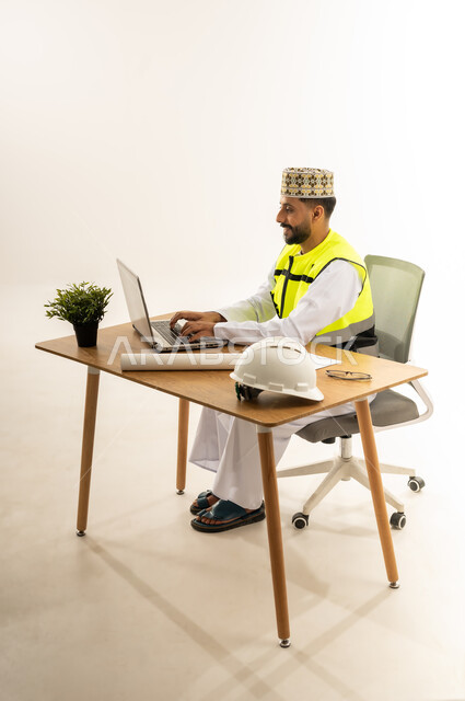 مفهوم إدارة المشاريع وتدقيقها، متابعة العمل ومراجعة مخطط المشروع، دمج التقنيات الحديثة بالقطاع الهندسي، بورتريه من الجانب لمهندس عربي خليجي عماني يرتدي الدشداشة مع سترة الحماية والكمة يجلس خلف المكتب أمامه الحاسوب المحمول وبجانبه النبتة والخوذة، خلفية بيضاء