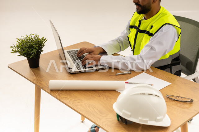 إدارة المشاريع بحرفية باستخدام الحاسوب المحمول، العمل في القطاع الهندسي، مفهوم الهندسة والانشاءات المعماريه، بورتريه مقرب من الجانب لمهندس عربي خليجي عماني يرتدي الدشداشة مع سترة الحماية يجلس خلف طاولة المكتب وأمامه المخطط والنبته وبجانبه الخوذة، خلفية بيضاء