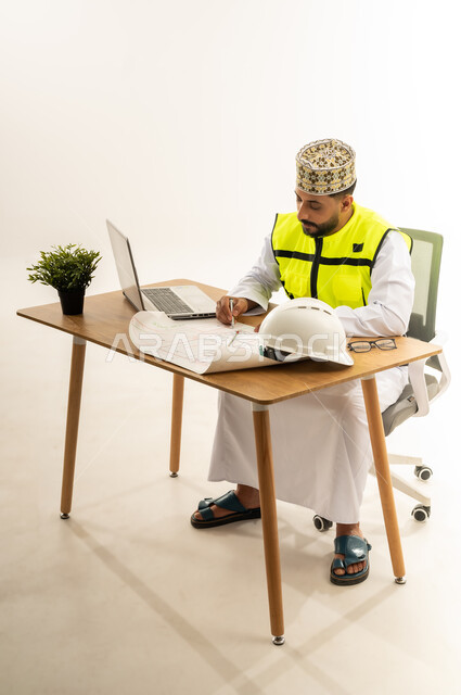 دمج التقنيات الحديثة بالقطاع الهندسي، مفهوم إدارة المشاريع وتدقيقها، بورتريه من الجانب لمهندس عربي خليجي عماني يرتدي الدشداشة مع سترة الحماية والكمة يجلس خلف المكتب أمامه الحاسوب المحمول وبجانبه النبتة والخوذة، متابعة العمل ومراجعة مخطط المشروع، خلفية بيضاء