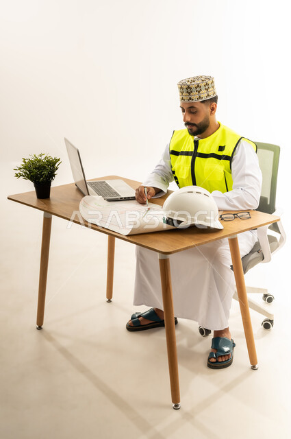 متابعة العمل ومراجعة مخطط المشروع، مفهوم إدارة المشاريع وتدقيقها، دمج التقنيات الحديثة بالقطاع الهندسي، بورتريه من الجانب لمهندس عربي خليجي عماني يرتدي الدشداشة مع سترة الحماية والكمة يجلس خلف المكتب أمامه الحاسوب المحمول وبجانبه النبتة والخوذة، خلفية بيضاء