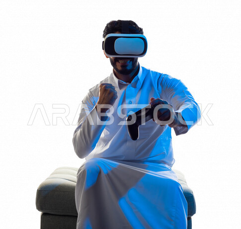 Portrait of a Saudi Arabian Gulf man wearing 3D virtual reality glasses, holding a joystick in his hand and playing video games with gestures indicating victory, fun and entertainment in virtual world games, PlayStation entertainment games, techniques and