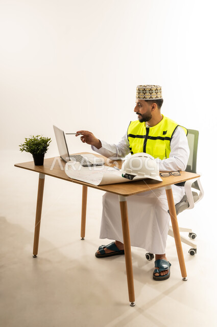العمل في القطاع الهندسي، مفهوم إدارة المشاريع وتدقيقها، بورتريه من الجانب لمهندس عربي خليجي عماني يرتدي الدشداشة مع سترة الحماية والكمة يجلس خلف طاولة المكتب وأمامه الحاسوب المحمول وبجانبه النبتة والخوذة، متابعة الاعمال ومراجعة مخطط المشروع، خلفية بيضاء