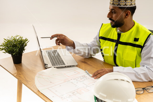Mastering project management using laptop, concept of engineering and architectural construction, working in engineering sector, close-up portrait from the side of an Arab Gulf Omani engineer wearing a dishdasha with a protective vest and a mask sitting behind an office table with a plan in front of him and a helmet next to him, white background