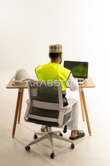 مراجعة مخطط المشروع ومتابعة الاعمال، العمل في القطاع الهندسي، مفهوم إدارة المشاريع وتدقيقها، بورتريه من الخلف لمهندس عربي خليجي عماني يرتدي الدشداشة مع سترة الحماية والكمة يجلس خلف طاولة المكتب وأمامه الحاسوب المحمول وبجانبه الخوذة، خلفية بيضاء