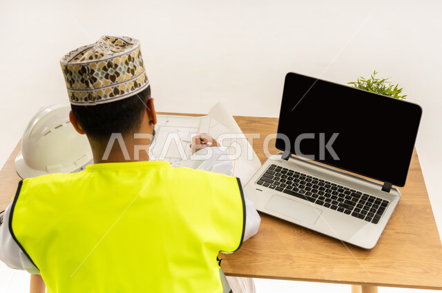 Concept of engineering and architectural construction, work in the engineering sector, concept of project management and auditing, top portrait from the back of an Arab Gulf Omani engineer wearing a dishdasha with a protective vest and a mask sitting behind an office table with a laptop in front of him and a helmet next to him, white background