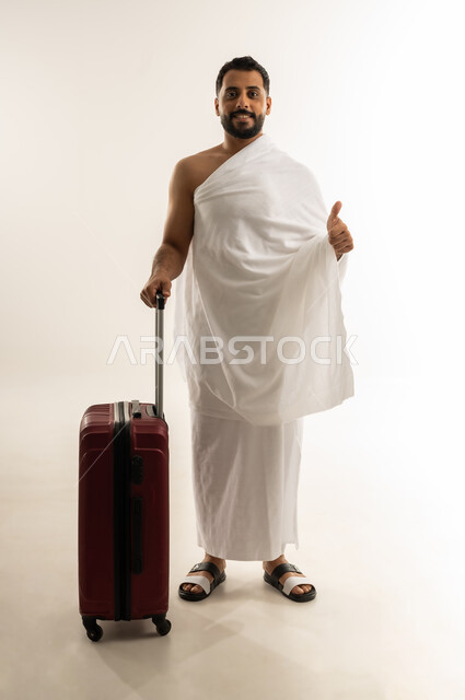 Thumb up sign, travel to perform Hajj and Umrah, worship, obedience and getting closer to Allah, portrait of an Arab Omani Gulf man wearing Ihram clothes and holding a red travel bag, looking at the camera with cheerful gestures, full body, white background