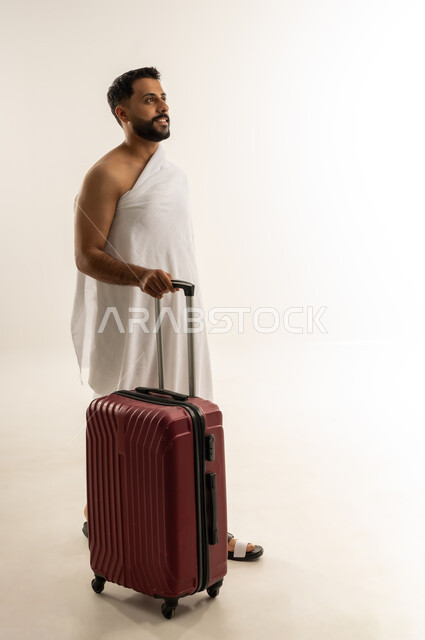 الاستعداد للسفر لأداء مناسك الحج والعمرة، العبادة والطاعة والتقرب إلى الله, بورتريه من الجانب لرجل عربي عماني خليجي يرتدي ملابس الإحرام ويجر حقيبة السفر الحمراء، النظر إلى الأعلى بإيماءات التأمل والتخطيط، الجسم بالطول الكامل، خلفية بيضاء