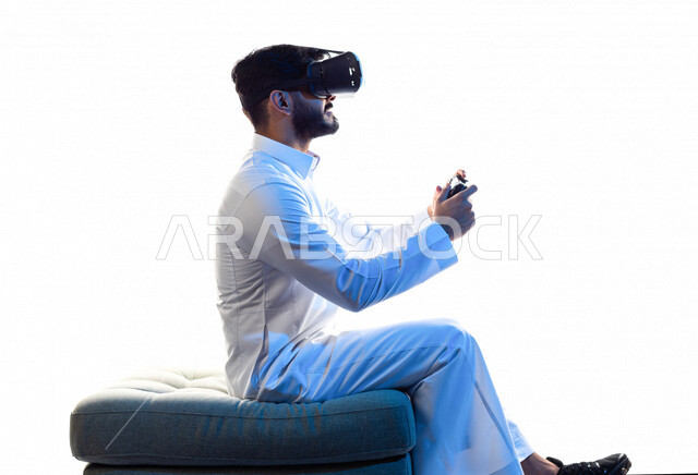 Portrait of a Saudi Arabian Gulf man wearing 3D virtual reality glasses, holding a joystick in his hand and playing video games with gestures indicating happiness, fun and entertainment in virtual world games, PlayStation entertainment games, technologies