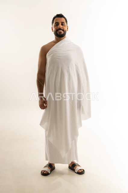 أداء مناسك الحج والعمرة، العبادة والطاعة والتقرب إلى الله, بورتريه لرجل عربي عماني خليجي يرتدي ملابس الإحرام ينظر إلى الكاميرا بإيماءات السرور والثقة بالنفس، الجسم بالطول الكامل، خلفية بيضاء