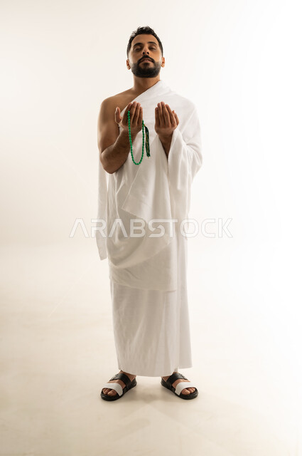 رفع الكفين نحو الأعلى والدعاء، طلب العفو والمغفرة من الله تعالى، العبادة والطاعة والإكثار من الأذكار, أداء مناسك الحج والعمرة، بورتريه لرجل عربي عماني خليجي يرتدي ملابس الإحرام يقف باستقامة ويدعو الله، الجسم بالطول الكامل، خلفية بيضاء