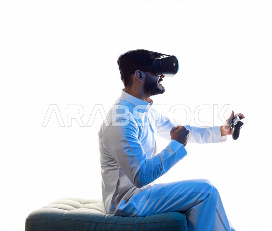 Portrait of a Saudi Arabian Gulf man wearing 3D virtual reality glasses, holding a joystick in his hand and playing video games with gestures indicating victory, fun and entertainment in virtual world games, PlayStation entertainment games, techniques and