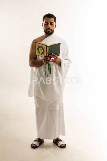 أداء مناسك الحج والعمرة، قراءة القرآن الكريم بتمعن وتركيز, تلاوة كتاب الله، بورتريه لرجل عربي عماني خليجي يرتدي ملابس الإحرام ويمسك بين يديه المصحف الشريف والسبحة، التقرب الى الله بأداء العبادة والطاعة, الجسم بالطول الكامل، خلفية بيضاء