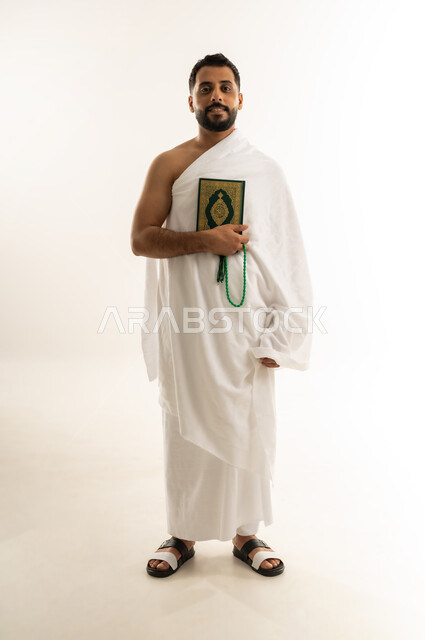 النظر إلى الكاميرا بإيماءات السعادة والسرور، التقرب الى الله بالعبادة والطاعة, أداء مناسك الحج والعمرة، قراءة القرآن الكريم, بورتريه لرجل عربي عماني خليجي يرتدي ملابس الإحرام ويمسك بين يديه المصحف الشريف والسبحة، الجسم بالطول الكامل، خلفية بيضاء