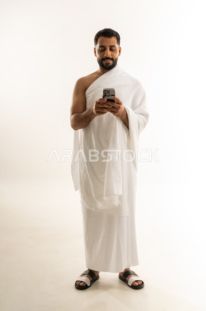 Benefiting from useful electronic applications via mobile phone, performing Hajj and Umrah rituals, worship, obedience and getting closer to God, portrait of an Arab Omani Gulf man wearing Ihram clothes holding a mobile device, electronic guide for Hajj and Umrah performers, full body length, white background