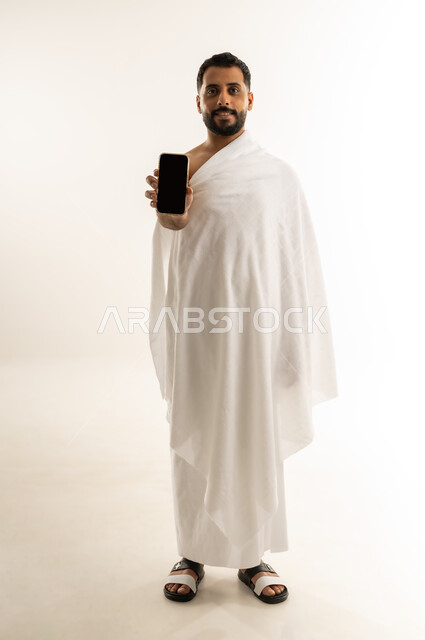 عرض شاشة هاتف محمول سوداء فارغة، أداء مناسك الحج والعمرة، العبادة والطاعة والتقرب إلى الله, بورتريه لرجل عربي عماني خليجي يرتدي ملابس الإحرام يمسك جهاز الجوال، النظر للكاميرا بإيماءات البشاشة، الجسم بالطول الكامل، خلفية بيضاء