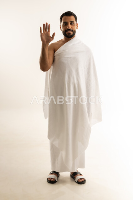 فتح كف اليد ورفعها نحو الأعلى بايماءات  السلام والوداع، أداء مناسك الحج والعمرة، العبادة والطاعة والتقرب إلى الله, بورتريه لرجل عربي عماني خليجي يرتدي ملابس الإحرام وينظر إلى الكاميرا بتعابير السرور والبشاشة، الجسم بالطول الكامل، خلفية بيضاء