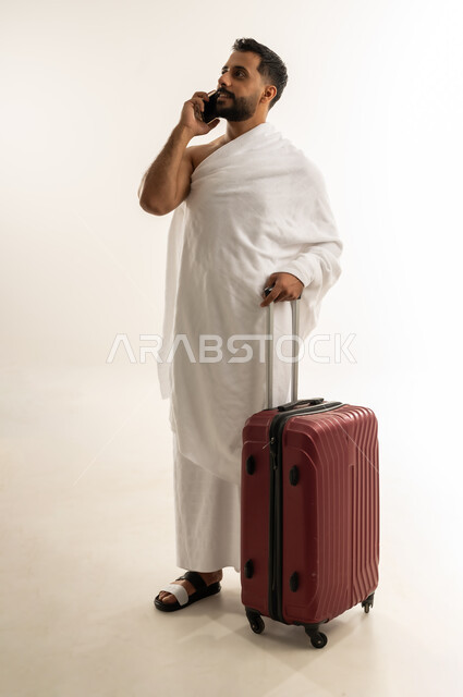 استخدام التقنيات التكنولوجية الحديثة، السفر إلى بيت الله الحرام لأداء مناسك الحج والعمرة، العبادة والطاعة والتقرب إلى الله, بورتريه لرجل عربي عماني خليجي يرتدي ملابس الإحرام ويمسك حقيبة السفر ويجري مكالمة هاتفية عبر الجوال، الجسم بالطول الكامل، خلفية بيضاء