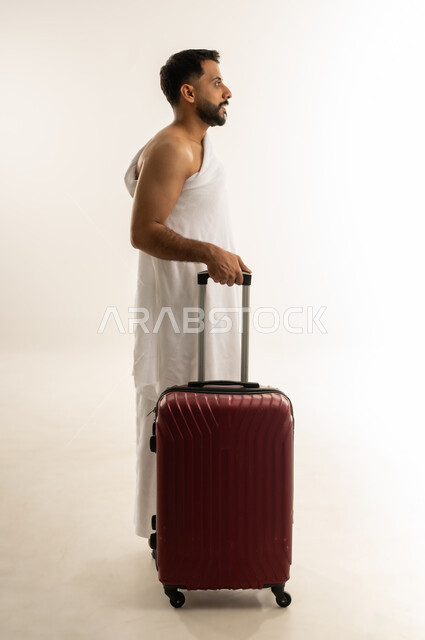 العبادة والطاعة والتقرب إلى الله, الاستعداد للسفر لأداء مناسك الحج والعمرة، بورتريه من الجانب لرجل عربي عماني خليجي يرتدي ملابس الإحرام ويمسك حقيبة السفر الحمراء، النظر إلى مكان ما بإيماءات التأمل والتخطيط، الجسم بالطول الكامل، خلفية بيضاء