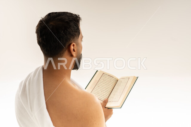 Reciting the Book of Allah, reading the Holy Quran with contemplation and focus, close-up portrait from the back of an Omani Gulf Arab man wearing Ihram clothes and holding the Holy Quran in his hands, performing the Hajj and Umrah rituals, getting closer to Allah by performing worship and obedience, white background