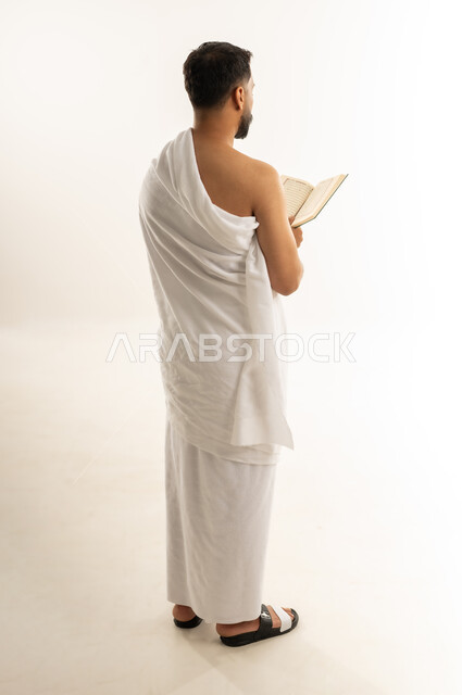 قراءة القرآن الكريم بتمعن وتركيز, تلاوة كتاب الله، بورتريه من الخلف لرجل عربي عماني خليجي يرتدي ملابس الإحرام ويمسك بين يديه المصحف الشريف، أداء مناسك الحج والعمرة، التقرب الى الله بأداء العبادة والطاعة, الجسم بالطول الكامل، خلفية بيضاء