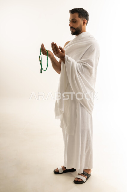 الخشوع والخضوع لله تعالى عند الدعاء، العبادة والطاعة والتقرب الى الله, التسبيح والتهليل وذكر الله، أداء مناسك الحج والعمرة، بورتريه من الجانب لرجل عربي عماني خليجي يرتدي ملابس الإحرام ويمسك السبحة بيده ويرفع كفيه نحو الأعلى، الجسم بالطول الكامل، خلفية بيضاء