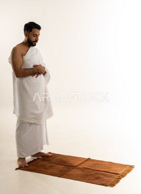 الوقوف على السجادة وأداء فريضة الصلاة، العبادة والطاعة والتقرب الى الله, مناسك الحج والعمرة، بورتريه من الجانب لرجل عربي عماني خليجي يرتدي ملابس الإحرام يتوجه للقبلة ويصلي، الجسم بالطول الكامل، خلفية بيضاء