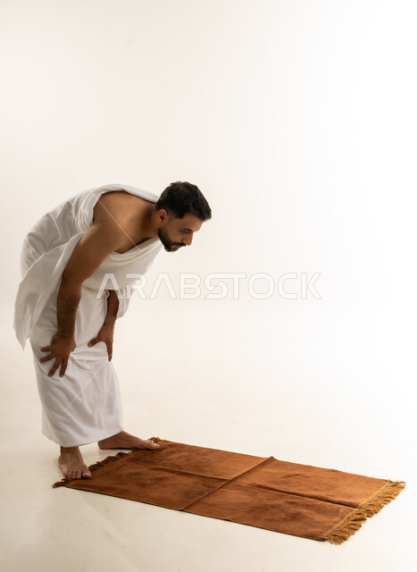 الركوع و أداء فريضة الصلاة، العبادة والطاعة والتقرب الى الله, مناسك الحج والعمرة، بورتريه من الجانب لرجل عربي عماني خليجي يرتدي ملابس الإحرام يتوجه للقبلة يقف على السجادة ويركع، خلفية بيضاء