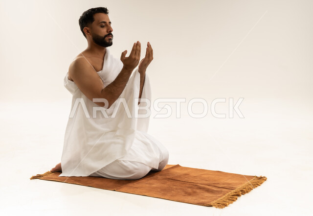 طلب المغفرة والعفو من الله، الدعاء بخشوع وتضرع، بورتريه لرجل عربي خليجي عماني يرتدي ملابس الإحرام يجلس على سجادة الصلاة يرفع كفيه المفتوحتين للأعلى ويدعو، أداء مناسك الحج والعمرة، التقرب إلى الله بالعبادة والطاعة، خلفية بيضاء